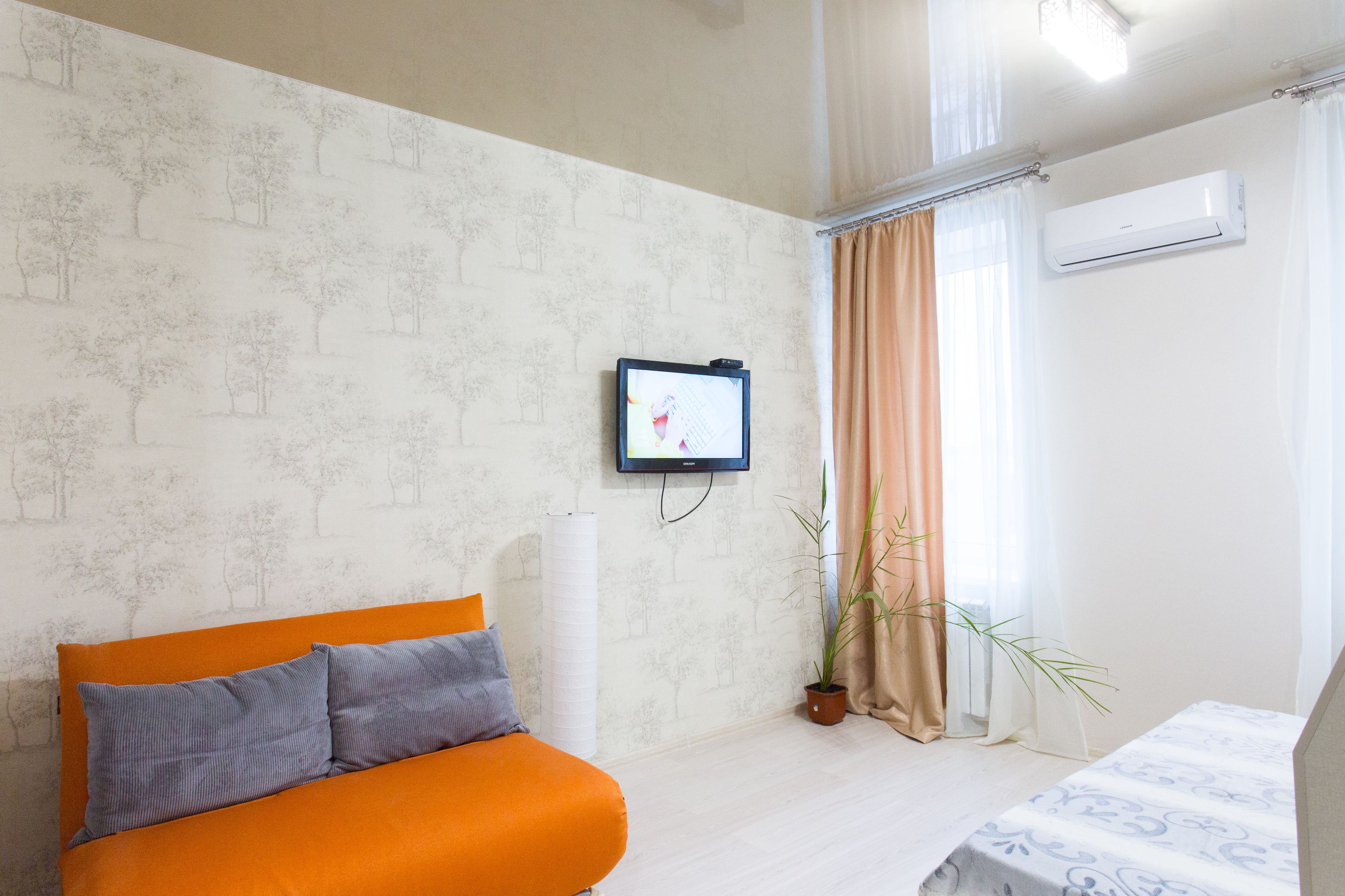 Una habitación con cama, sofá con cojines, televisor montado en la pared, planta en maceta y ventana con cortinas.