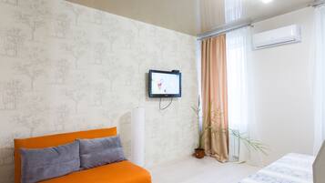 Una habitación con cama, sofá con cojines, televisor montado en la pared, planta en maceta y ventana con cortinas.