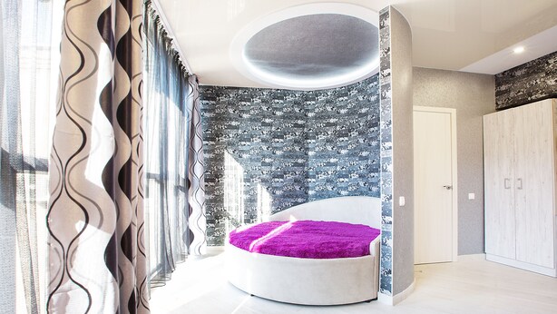 A modern bedroom with a circular bed, a large window with curtains, and a view of the cityscape.