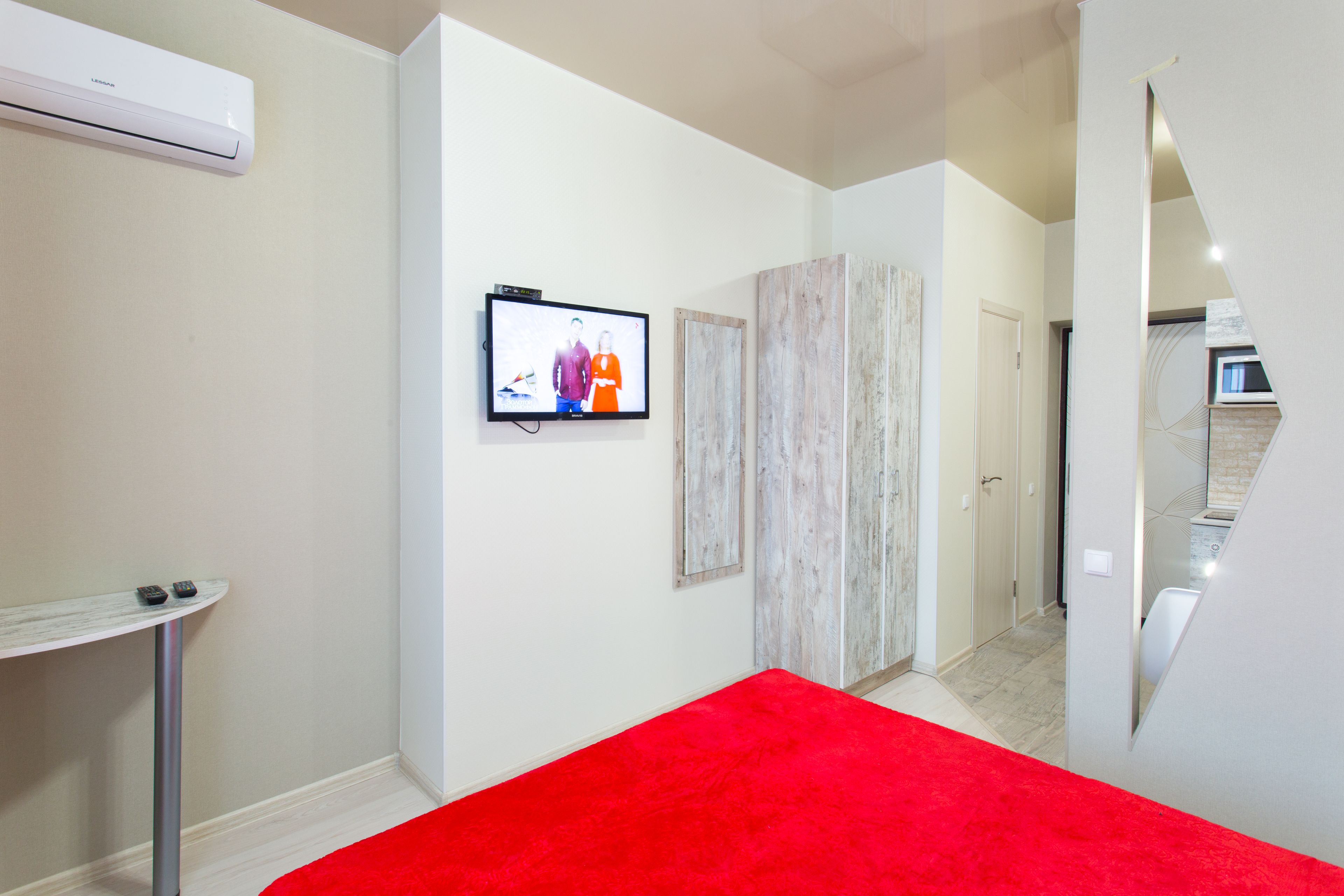 A modern hotel room with a red carpet, a mounted TV, a small table with remotes, and a wooden wardrobe.