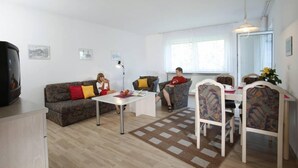 A modern living room with a sofa, a dining table, and chairs. There is a television on a stand and a window providing natural light.