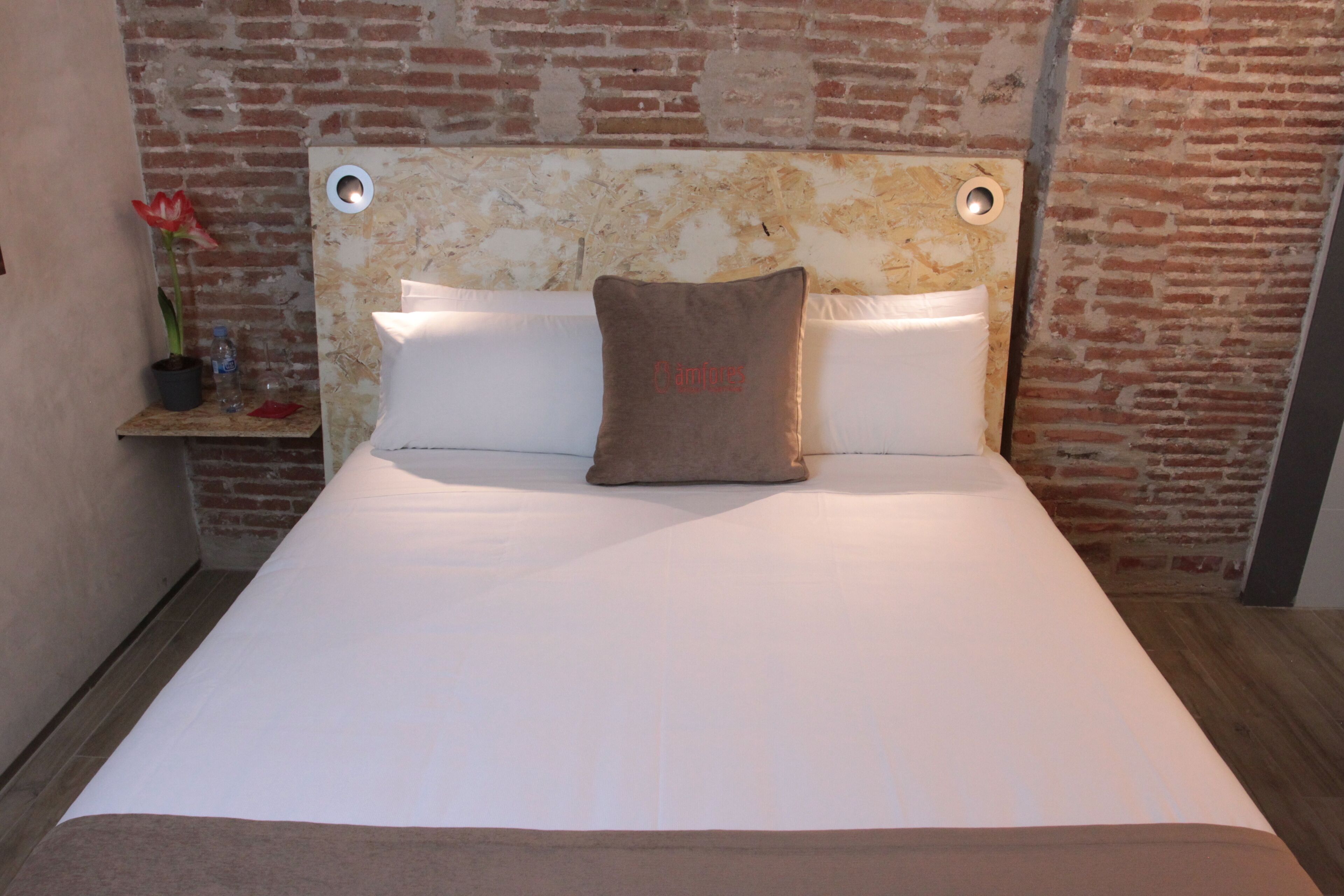 A bed with a headboard against a brick wall, a bedside table with a potted plant, and a pillow with the word "Cambridge" on it.