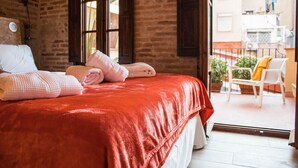 A hotel room with a brick wall, a large bed with a red blanket, a white chair, and a balcony with plants. - Amfores Boutique Guest House (Barcelona)