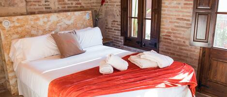 A bed with white linens and a red blanket, a wooden headboard, and a brick wall.