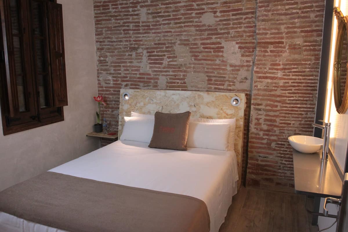 Ein Hotelzimmer mit Backsteinwand, einem Bett mit weißer Bettwäsche, einem Nachttisch mit Lampe und einem Badezimmer mit Waschbecken und Spiegel.