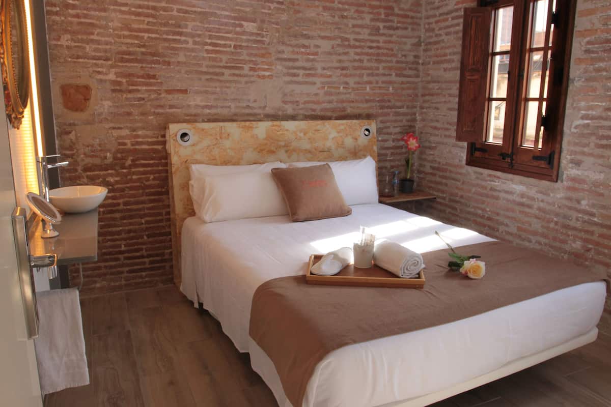 Ein Hotelzimmer mit Backsteinwand, einem hölzernen Kopfteil, einem Bett mit weißen Leinen, einem Tablett mit Handtüchern und Pflegeprodukten sowie einem Fenster mit Holzjalousien.