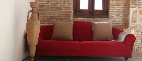 A red sofa with two brown cushions against a brick wall, a wooden floor, and a window with a wooden frame.