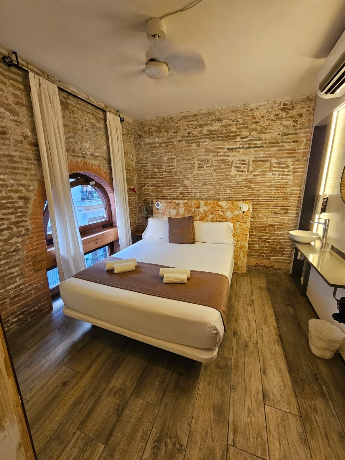 Un dormitorio con pared de ladrillo, suelo de madera, cama con ropa blanca, mesita de noche y ventana con cortinas.