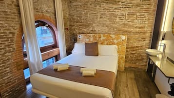 Een slaapkamer met een bakstenen muur, een houten vloer, een bed met witte lakens, een nachtkastje en een raam met gordijnen.