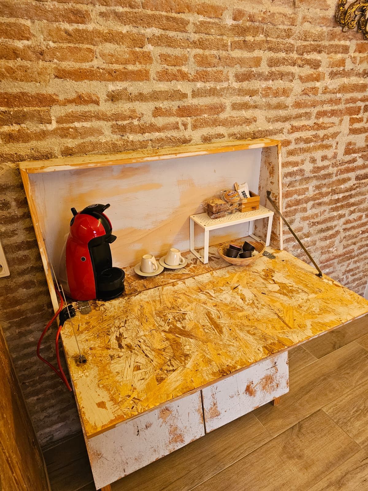 A rustic wooden table with a coffee machine and cups, set against a brick wall.