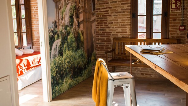 A room with a wooden dining table, a white stool with a yellow towel, a mural of a waterfall, and brick walls. - Amfores Boutique Guest House (Barcelona)