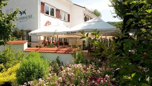Un hotel con terraza al aire libre bajo sombrillas, rodeado de jardines exuberantes y plantas con flores.