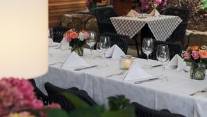 Un área de comedor al aire libre con mesas dispuestas para dos, rodeada de sillas y un muro de madera al fondo.