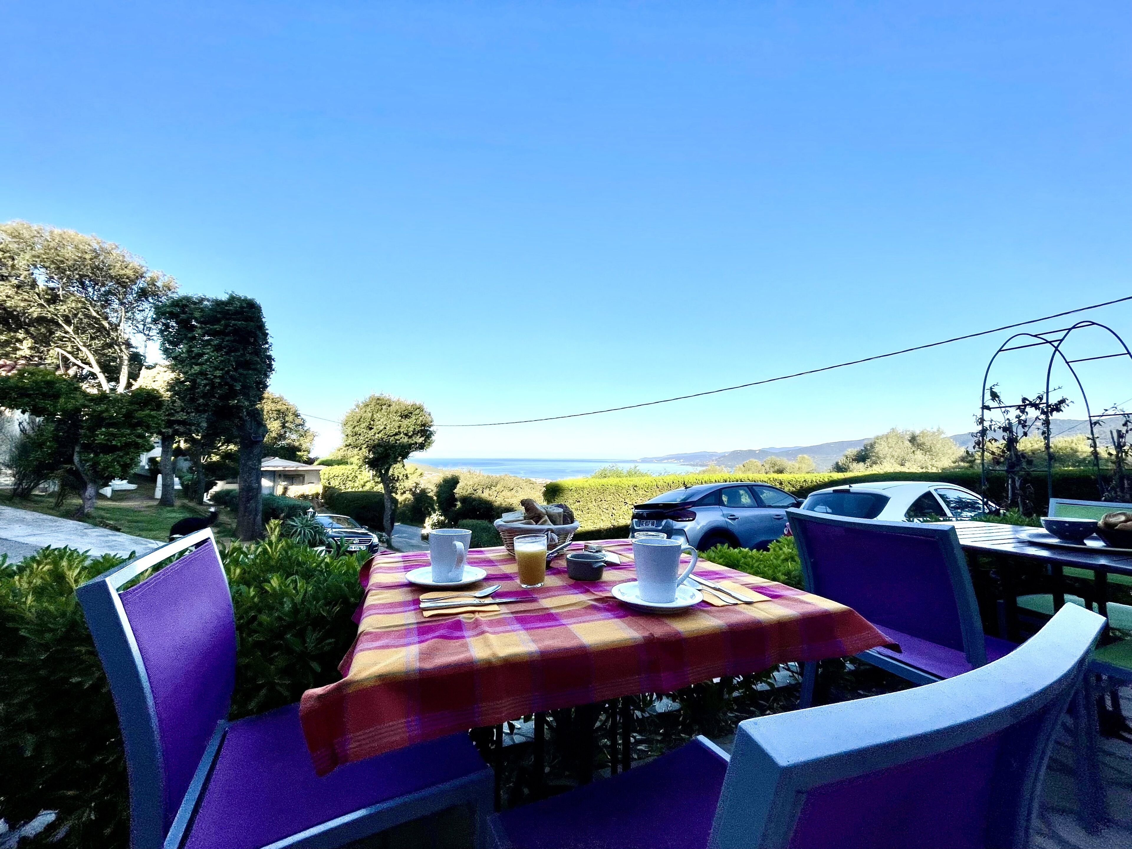Un espace de restauration en plein air avec une table dressée pour deux personnes, offrant une vue sur un paysage pittoresque où sont garées quelques voitures à proximité.