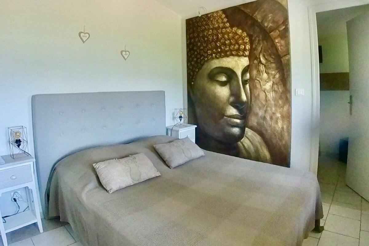 Een slaapkamer met een groot boeddhabeeld aan de muur, een bed met twee kussens, een nachtkastje en een wandlamp.