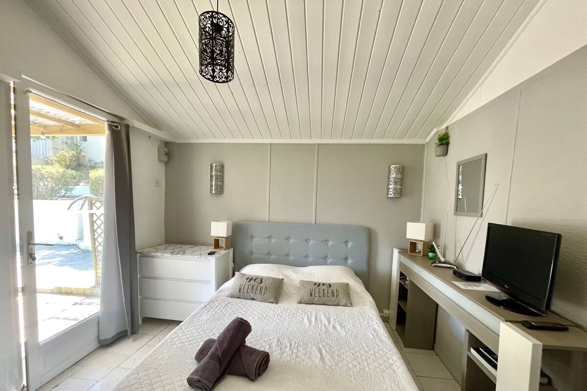 Een slaapkamer met een bed, nachtkastjes, een bureau met een televisie en een deur naar een balkon.