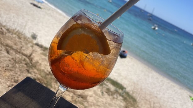 Een glas amberkleurig drankje op een houten tafel, met een strand en boten op de achtergrond.