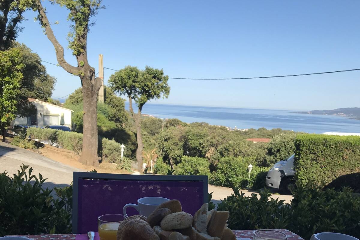 Een ontbijttafel met brood, sap en koffiekopjes, met uitzicht op een prachtig zeegezicht en groen.