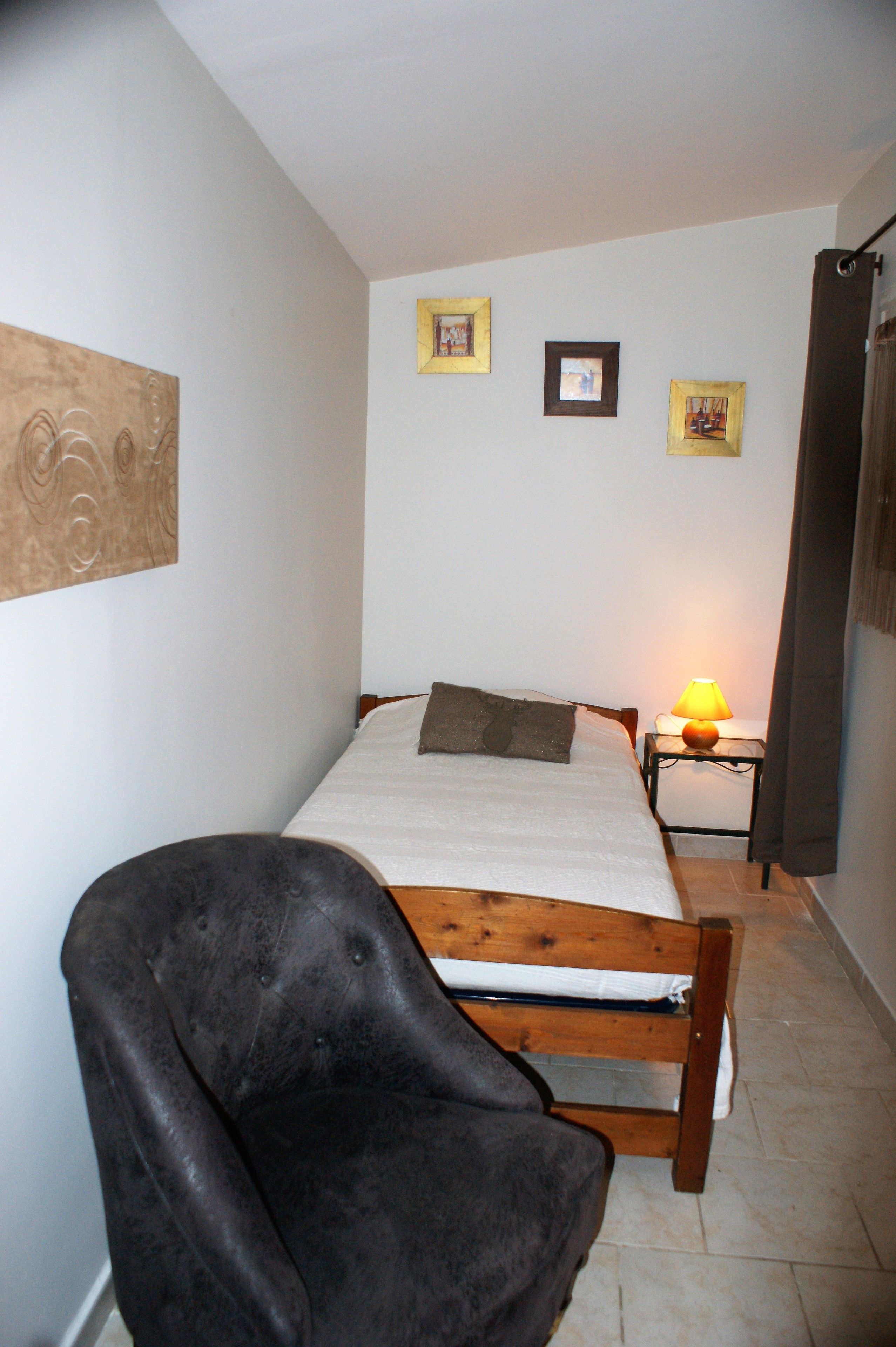 A small, narrow room with a single bed, a chair, and a nightstand with a lamp. There are three framed pictures on the wall.