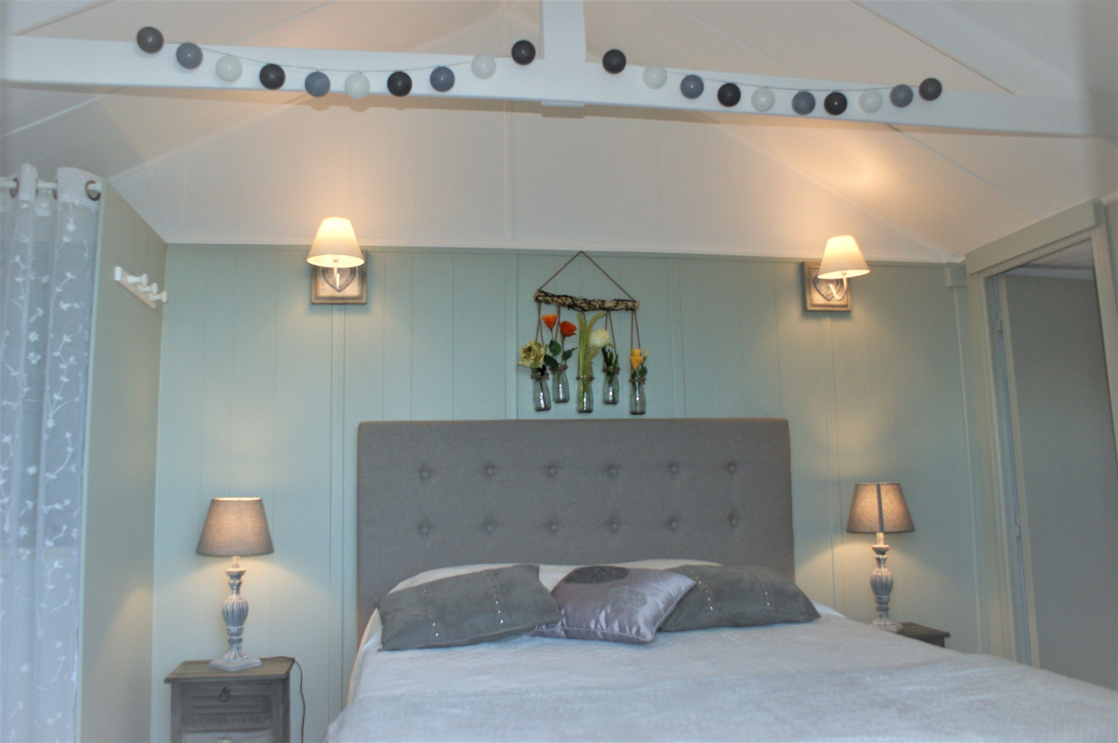 Un dormitorio con cama, dos mesitas de noche, dos lámparas, un arreglo floral colgante y una cortina.