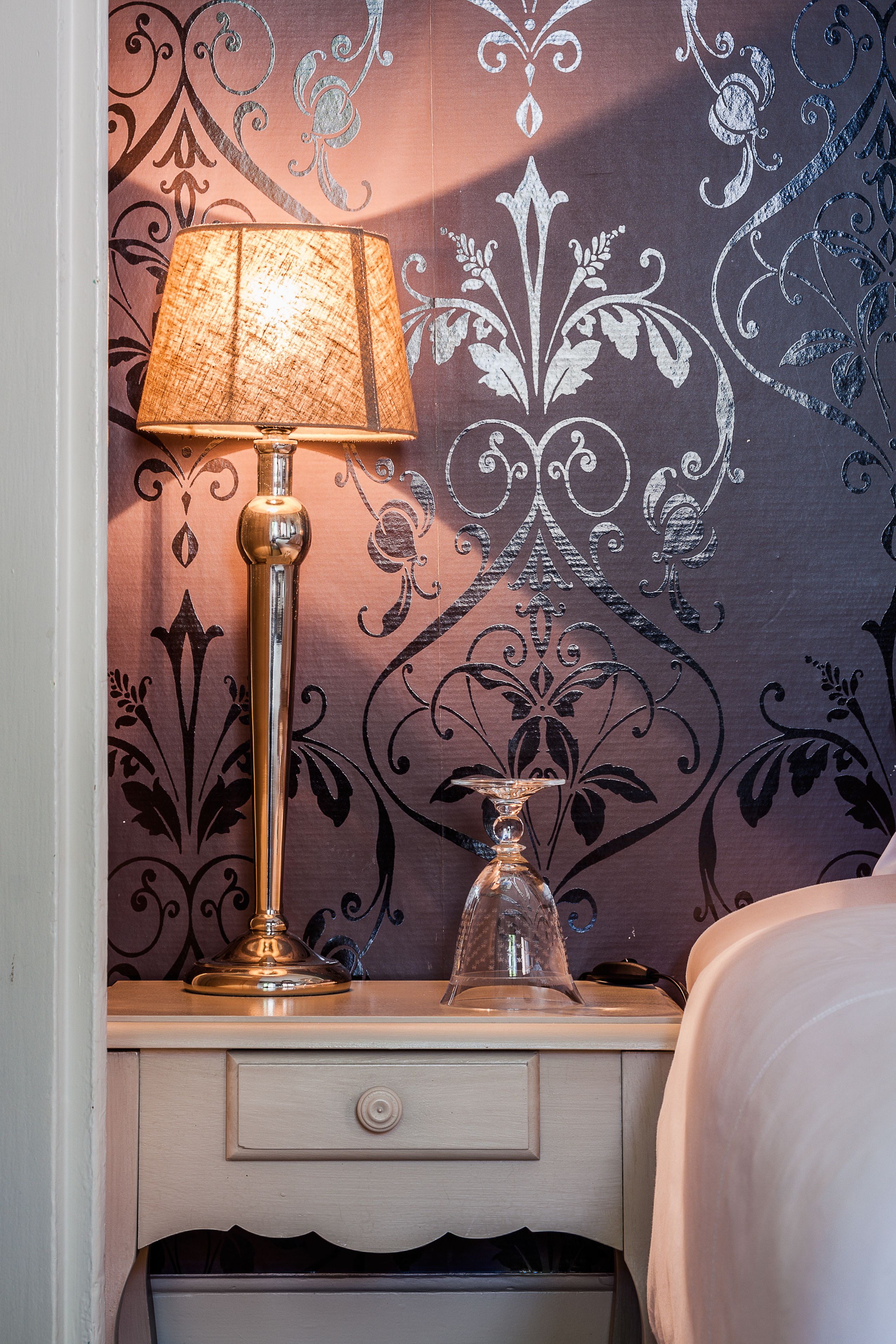 A bedside table with a lamp, a decorative glass vase, and a patterned wallpaper.
