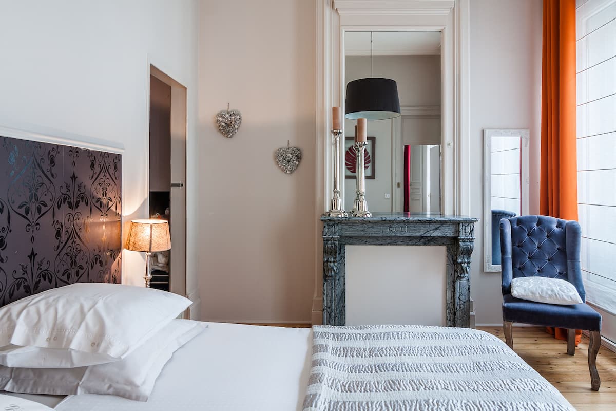 A bedroom with a bed, a nightstand, a dresser, a mirror, a fireplace, and a window with curtains.