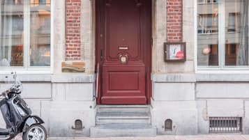 Une porte rouge avec un heurtoir en laiton, un scooter garé sur le côté gauche et une enseigne accrochée au mur, à droite.