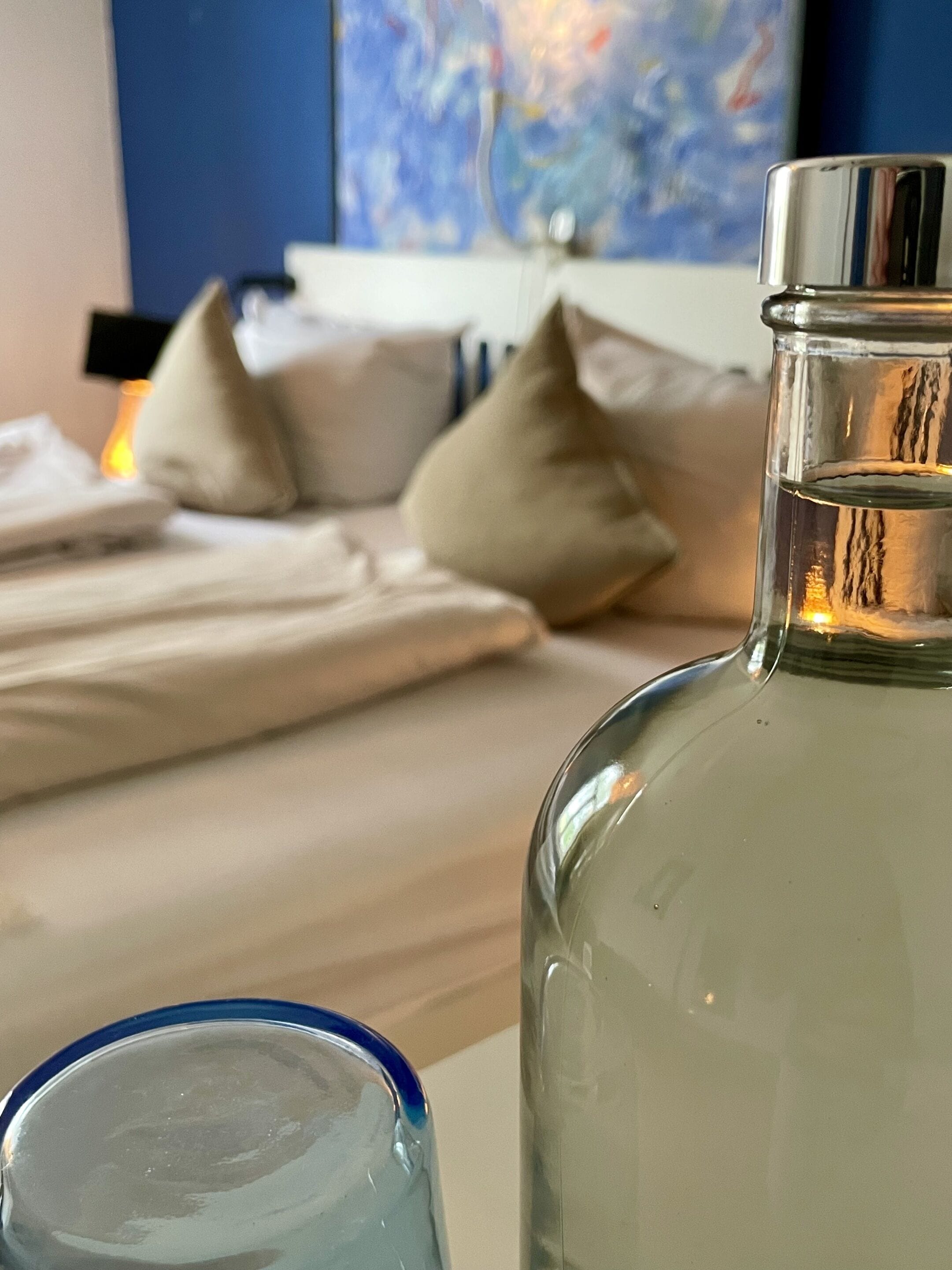 A clear glass bottle on a bedside table with a blue rim, a white pillow, and a bed with white bedding.