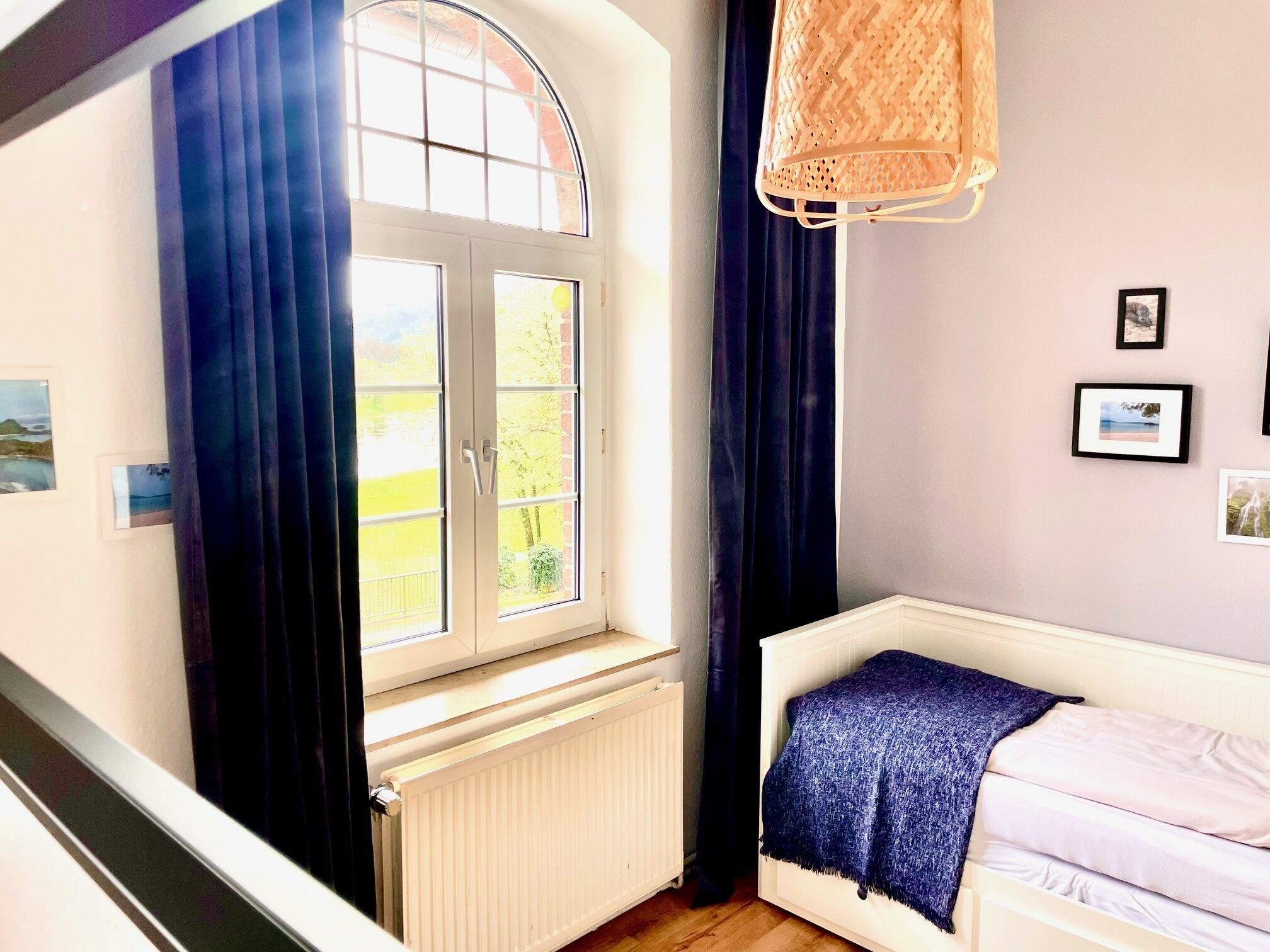 A bedroom with a bed, a window with curtains, a radiator, and framed pictures on the wall.