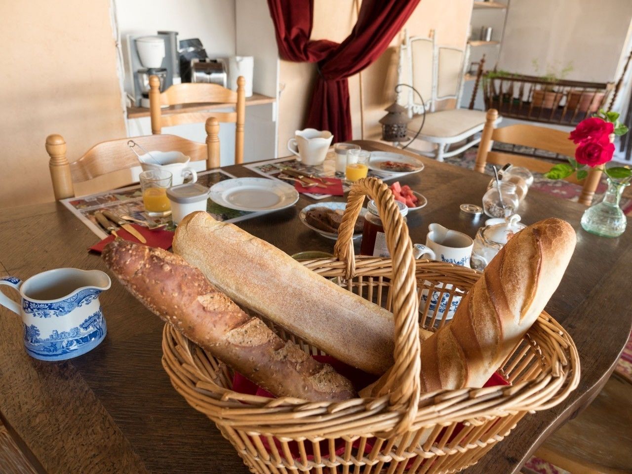 Ein Esstisch mit einem Korbwäscher, der Baguettes, eine blau-weiße Keramikkaraffe und ein Glas Orangensaft enthält.