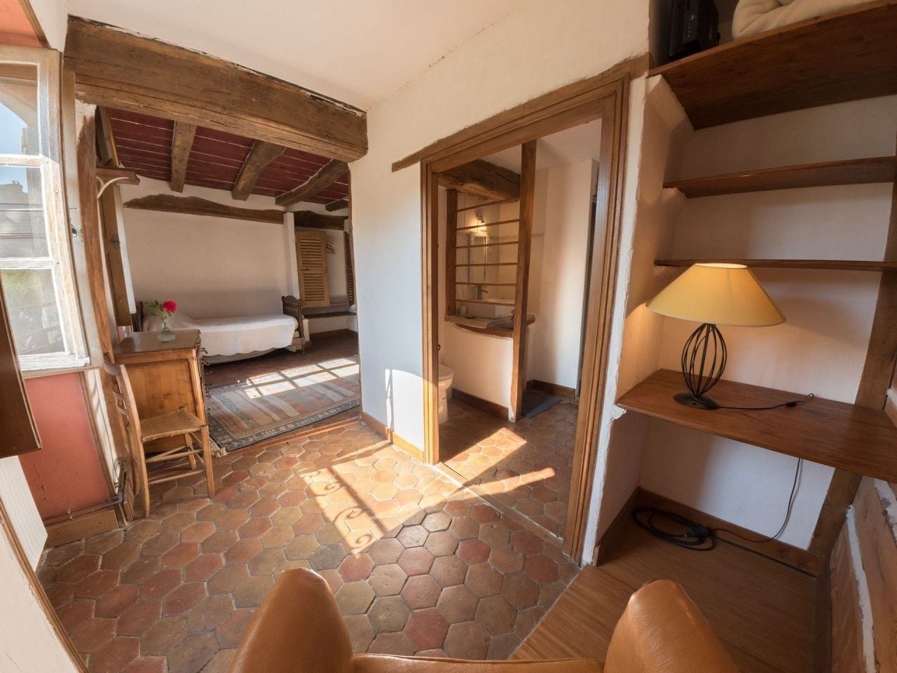 A cozy room with a bed, a wooden chair, a small table, and a shelf with a lamp.