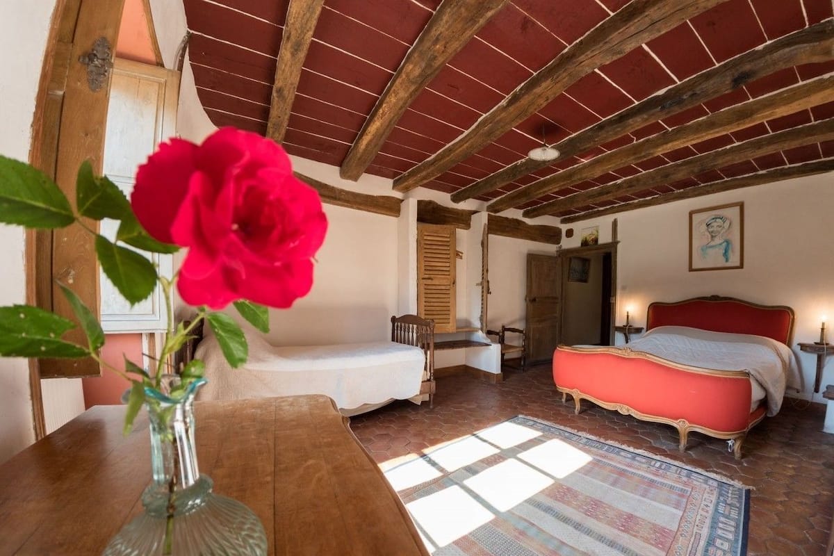 Quarto com teto de madeira, cama vermelha, banco de madeira e um vaso com uma flor vermelha.