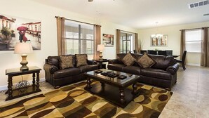 Un salon avec deux canapés en cuir marron foncé, une table basse en verre et un tapis à motifs. On y trouve également un ventilateur de plafond, un coin repas avec une table et des chaises, ainsi qu’un tableau accroché au mur.