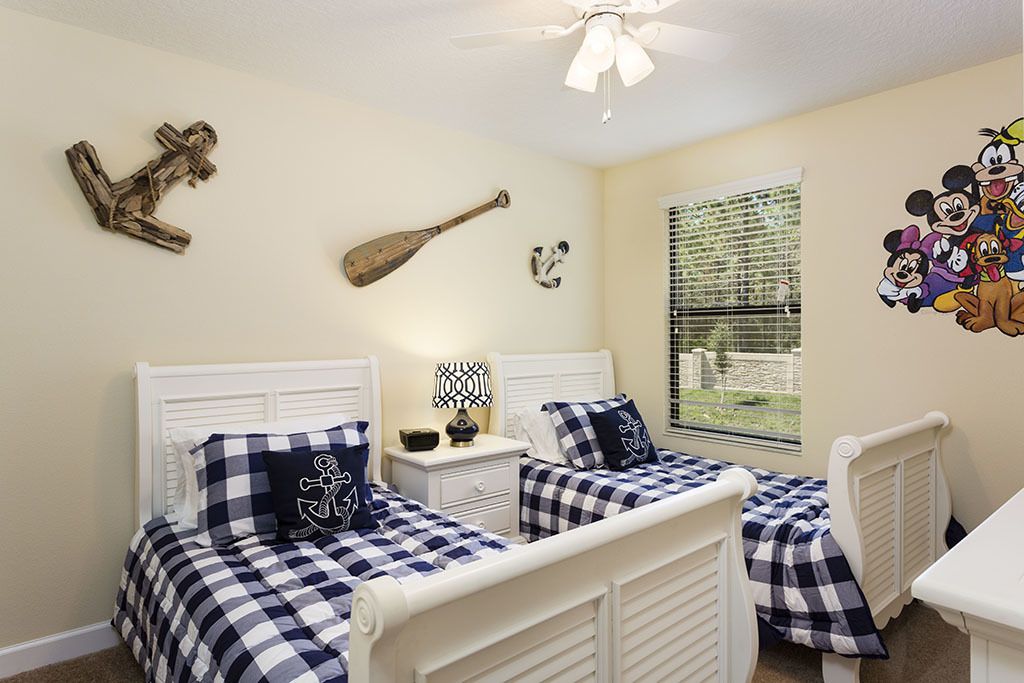 A room with two beds, a ceiling fan, a window with blinds, and wall decorations including a paddle, a wall-mounted clock, and a Mickey Mouse mural.