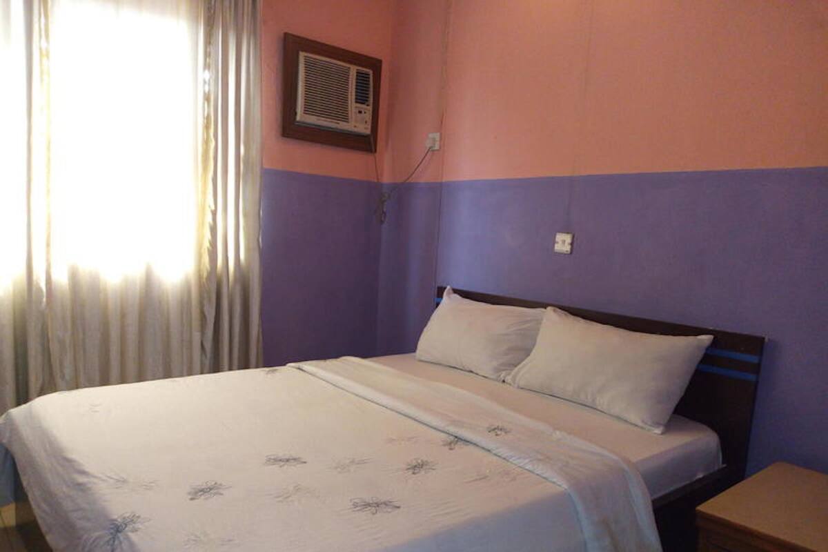 Una cama individual con una colcha blanca y dos almohadas en una habitación con paredes moradas y una ventana con cortinas.