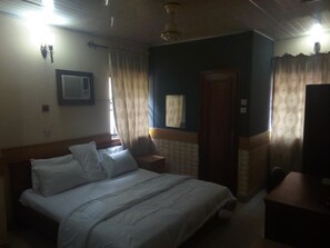 A bedroom with a bed, a nightstand, a wooden headboard, a ceiling fan, and a window with curtains. - Holla-Jobi Guest House (Lagos)