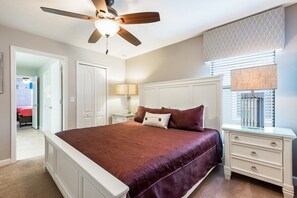 A bedroom with a large bed, a ceiling fan, a nightstand, and a window with blinds.