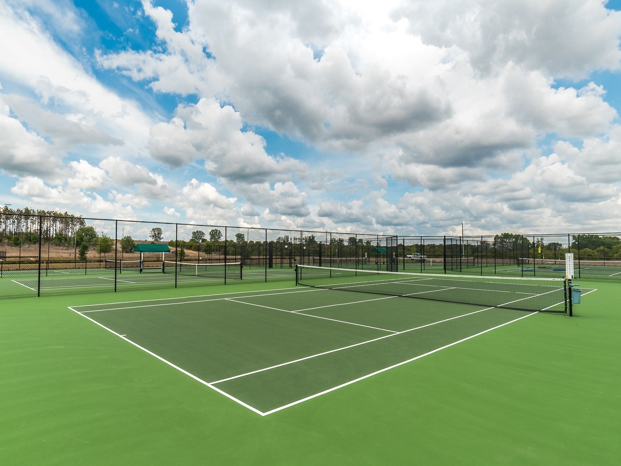 A tennis court with a green surface and white boundary lines, surrounded by a black fence, under a partly cloudy sky.