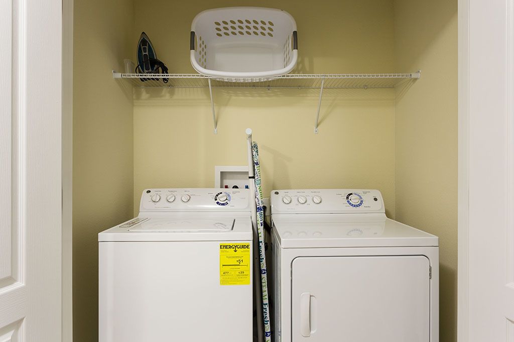 A laundry room with two white front-loading washing machines, a white basket on a shelf, and a wall-mounted dryer.