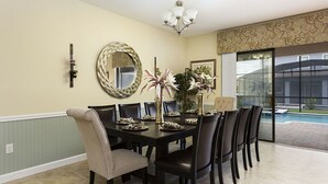 Une salle à manger avec une grande table, des chaises, un lustre, un miroir décoratif et une vue sur une piscine.