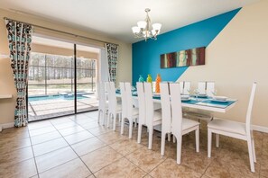 Uma sala de jantar com uma mesa grande posta para seis pessoas, cadeiras brancas, uma parede azul e vista para uma piscina através de portas de vidro.