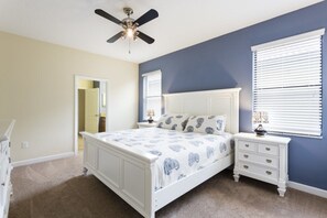 Une chambre à coucher comprenant un lit, des tables de chevet, un ventilateur de plafond et une porte donnant sur une autre pièce.
