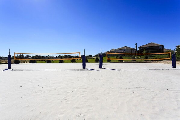 An outdoor volleyball court with a net, surrounded by sand and houses in the background.
