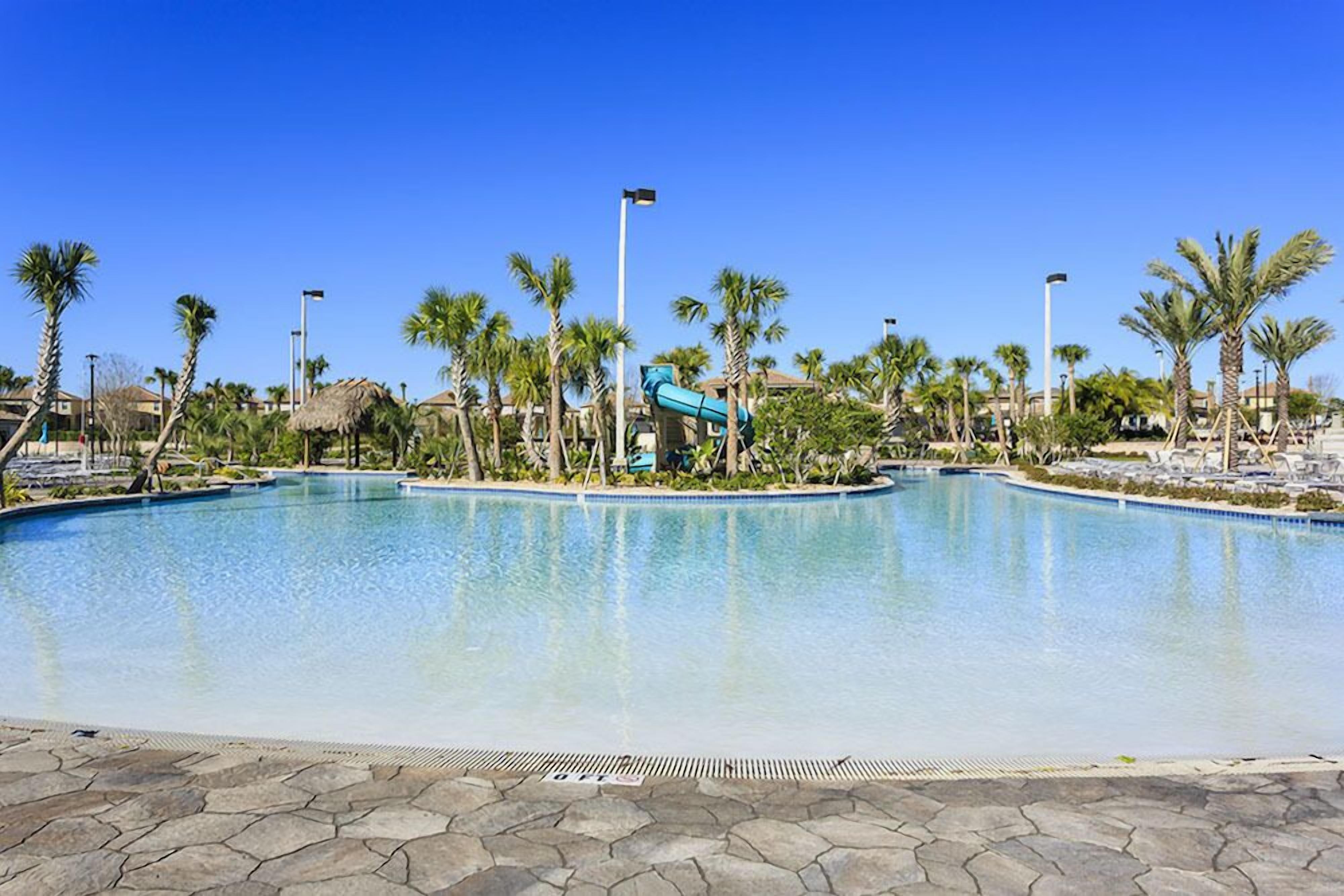 A large pool with a water slide, surrounded by palm trees and a clear blue sky.