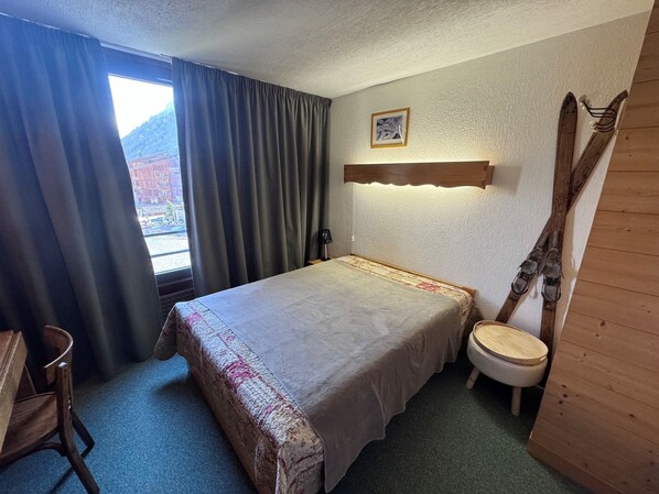 A hotel room with a bed, a wooden chair, a small table, a wooden bench, and a view of the outside.