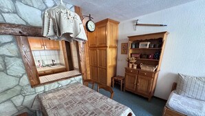 A room with a stone wall, wooden cabinets, a table with a patterned tablecloth, a bed with a white mattress, and a clock on the wall.