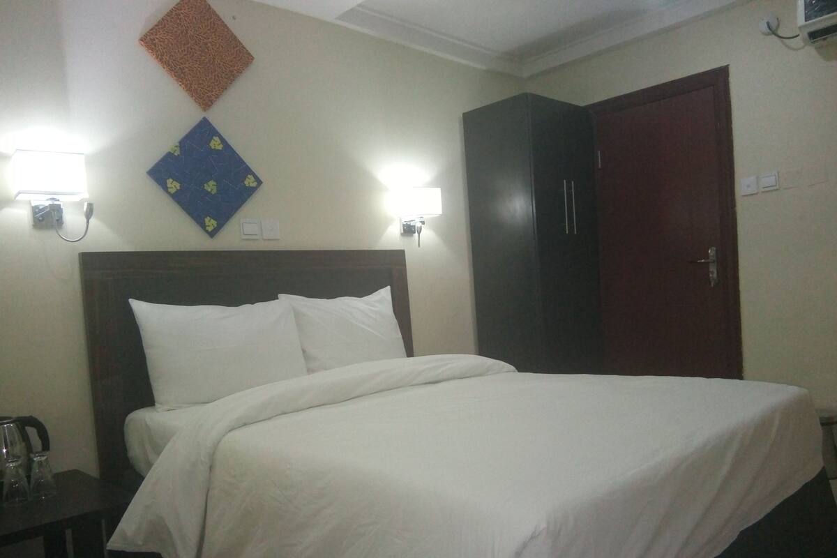 Hotellihuone, jossa on sänky, vaatekaappi ja yöpöytä, jossa on lasi.