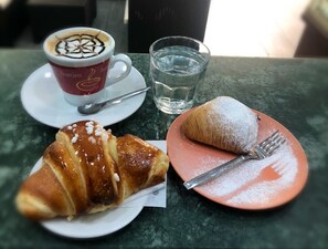 Un croissant, una taza de café con arte de latte y un vaso de agua sobre una mesa.