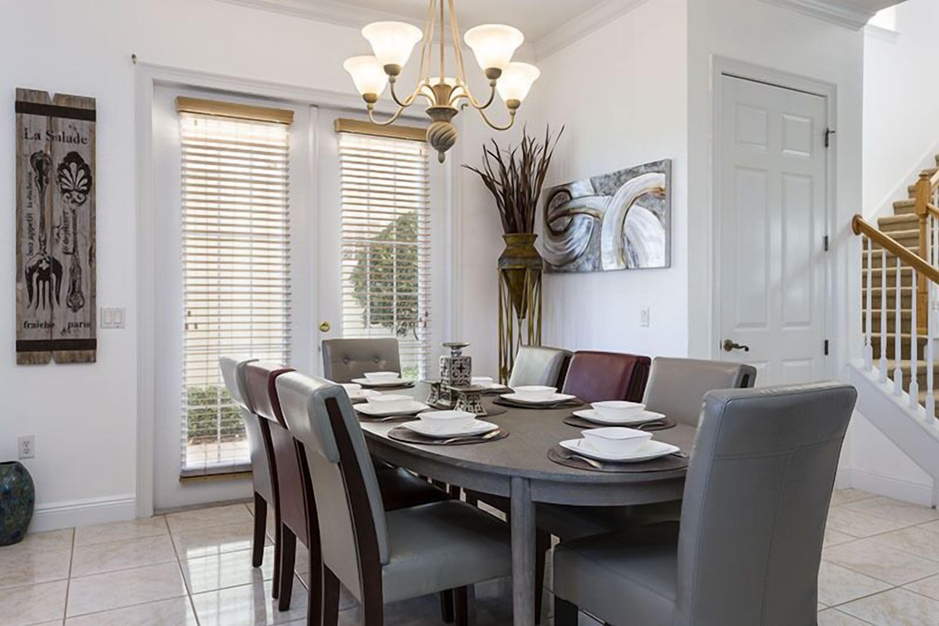 A dining room with a round table set for six, surrounded by chairs. There is a chandelier above the table, and a vase with plants on the wall.
