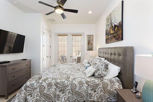 A bedroom with a bed, a television, a ceiling fan, a painting, and a window with blinds.
